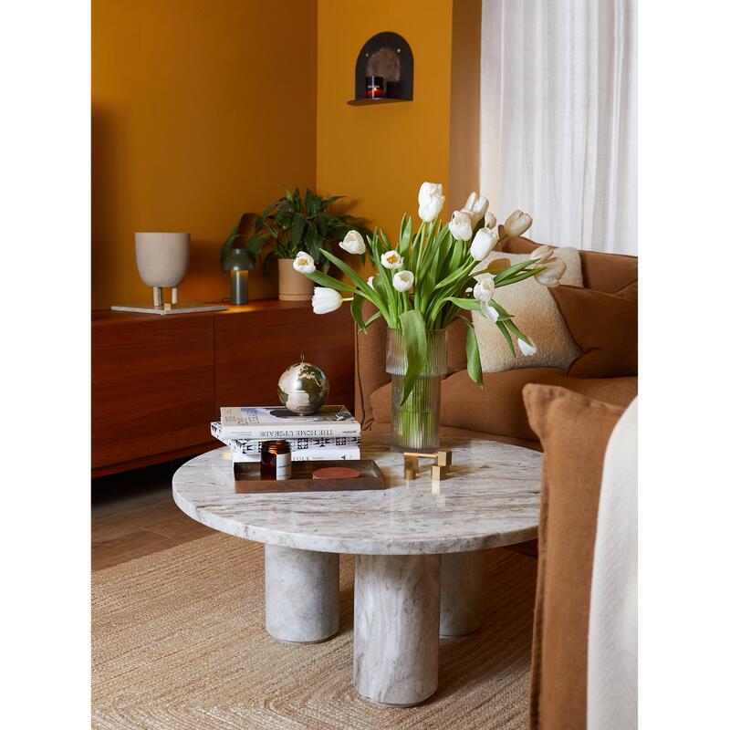 A vase of white tulips sits on a marble coffee table surrounded by books and a small globe, in a warm, mustard-yellow living room with wooden cabinets, plants, and cozy seating.