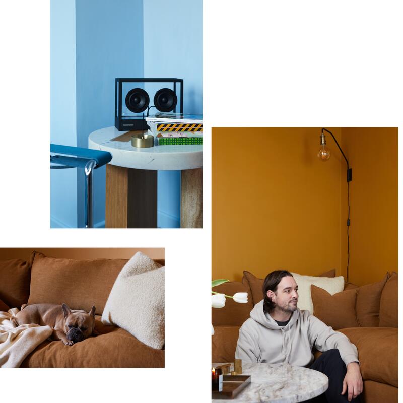 A round table with books and speakers, a wall-mounted light bulb, and a man sitting on a brown sofa with a white pillow. A dog sleeps on the same sofa.