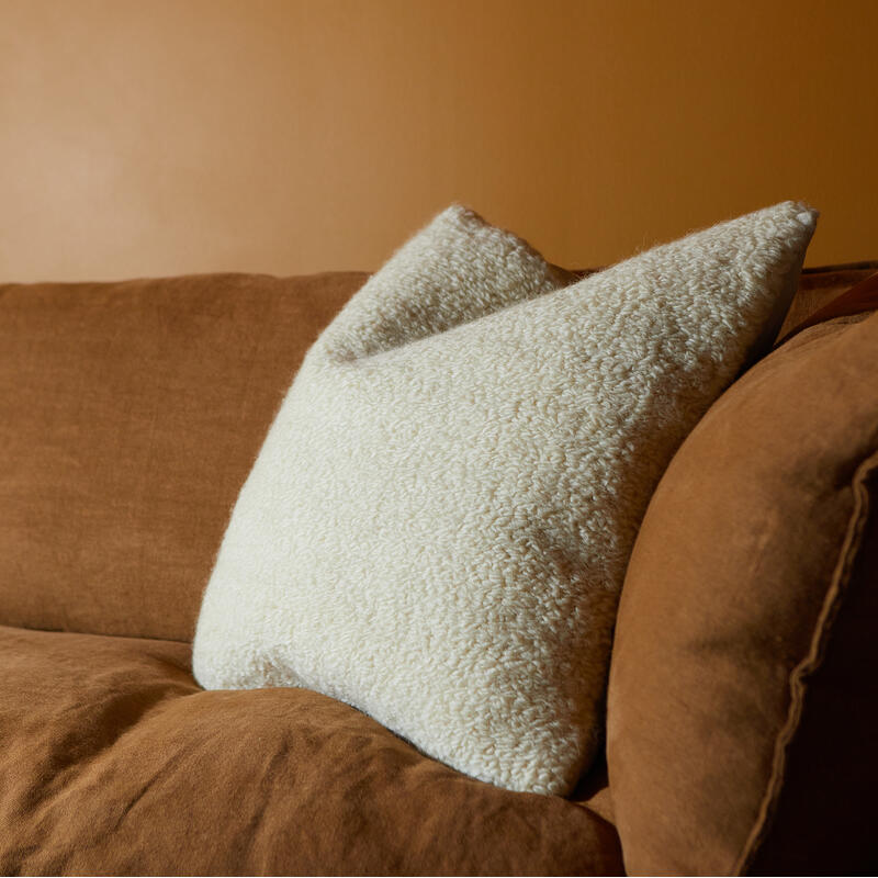 A beige, textured cushion rests on a brown sofa against an orange wall, providing a warm, cozy atmosphere.