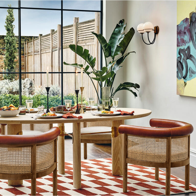 Dining table set with food and glasses, surrounded by wicker chairs, situated on a red-and-white checkered rug. The backdrop features large windows showing a garden with tall trees.