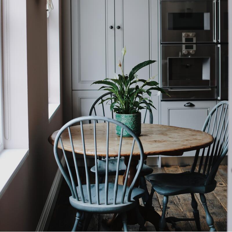 A potted plant sits on a round wooden table with four blue chairs. The table is by a window, in a kitchen with grey cabinets and built-in appliances.