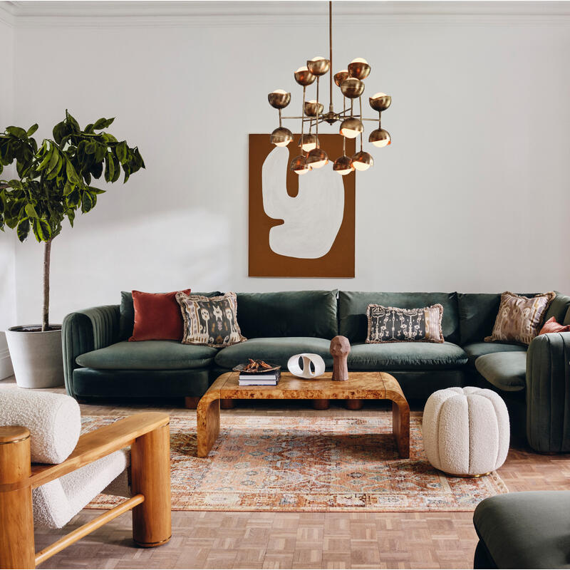 A large green sectional sofa with various cushions occupies a modern living room with wooden furniture, abstract art, a potted plant, a floor lamp, and large windows, creating a cozy ambiance.