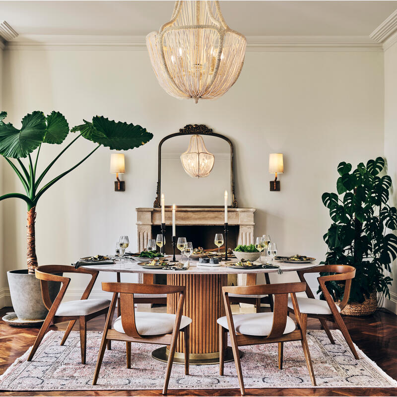 Dining table set with plates and glasses, surrounded by four chairs, situated in a stylish room with large houseplants, a chandelier, a fireplace with candles, and a rug underneath.