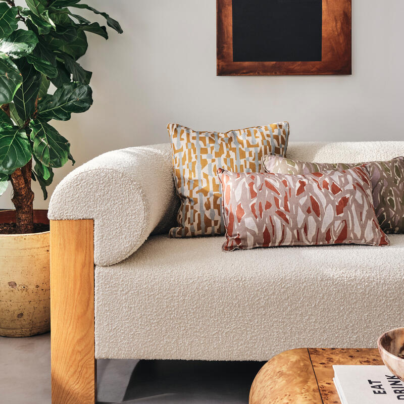 Beige textured sofa adorned with patterned cushions is placed beside a large potted green plant, with a wooden coffee table holding a book titled "EAT DRINK" and ceramic bowl nearby.