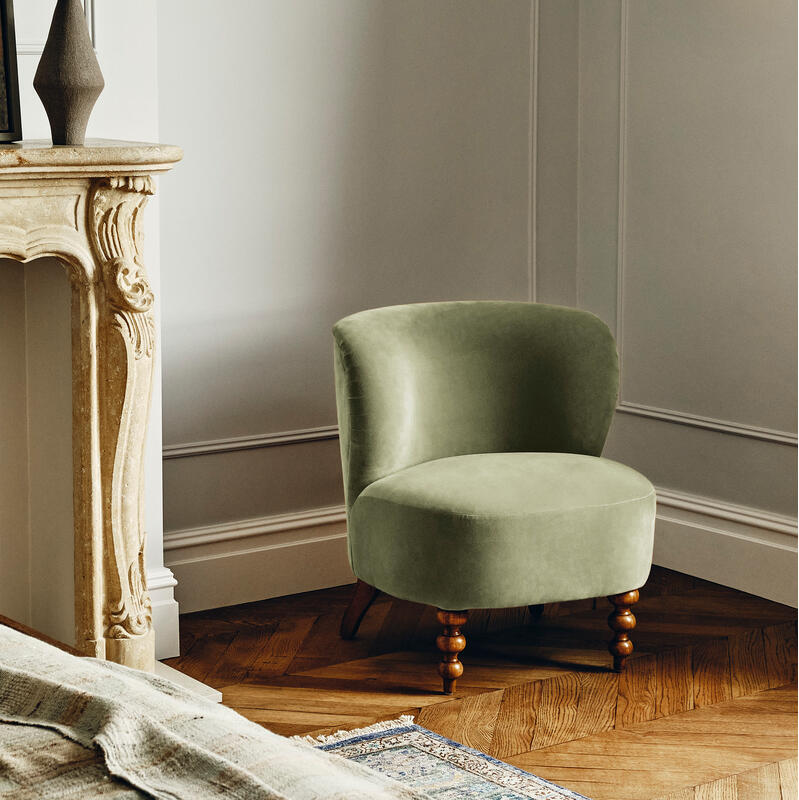 A green upholstered chair sits on wooden herringbone flooring near an ornate fireplace with a framed abstract painting and vase, alongside a bed draped with a textured blanket in a cozy room.