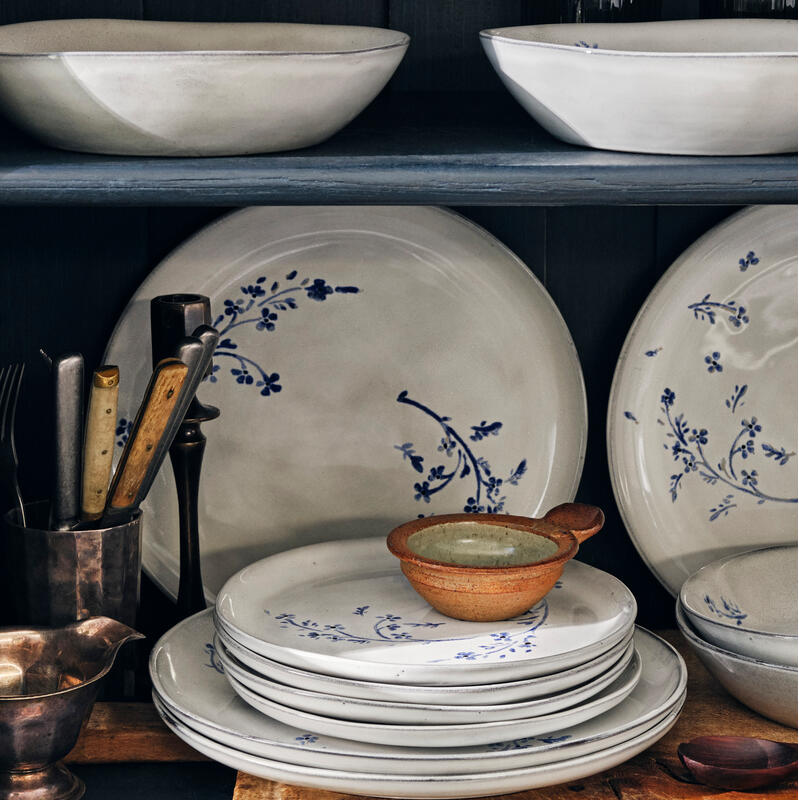 Stacked white plates with blue floral patterns, placed on a rustic wooden shelf along with vintage utensils and bowls, inside a dark-colored wooden cabinet.