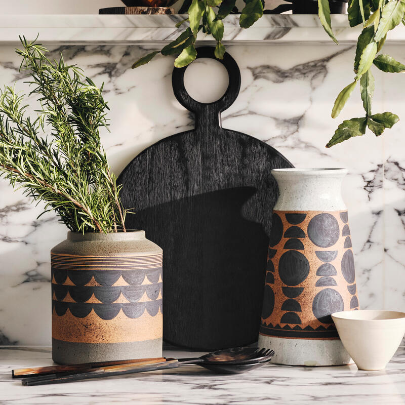 Ceramic jars holding green plants and cutlery rest on a marble countertop; a black cutting board stands behind them, set against a white wall with a shelf displaying a gold cup and potted plant.