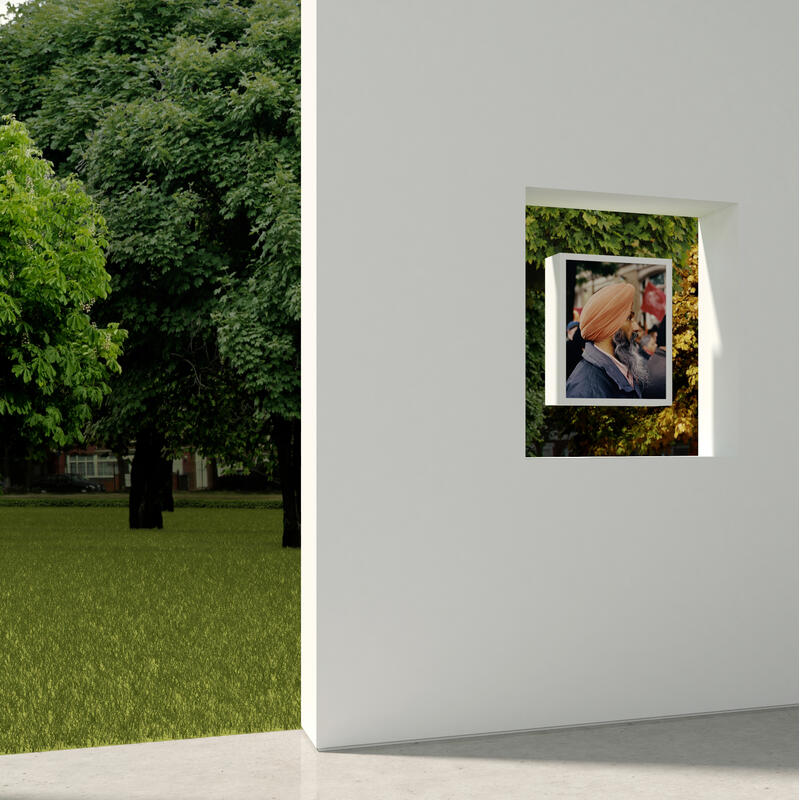 Framed photograph of a man with a turban and beard hanging on a white wall, with another framed portrait leaning against the wall, and a lush green park visible through a window.