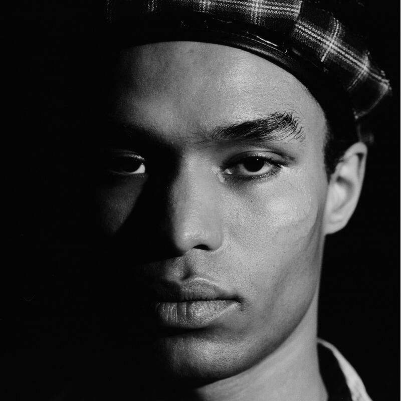 A young person wearing a checkered beret and leather jacket gazes seriously into the camera, with dramatic shadows accentuating their face against a dark background.