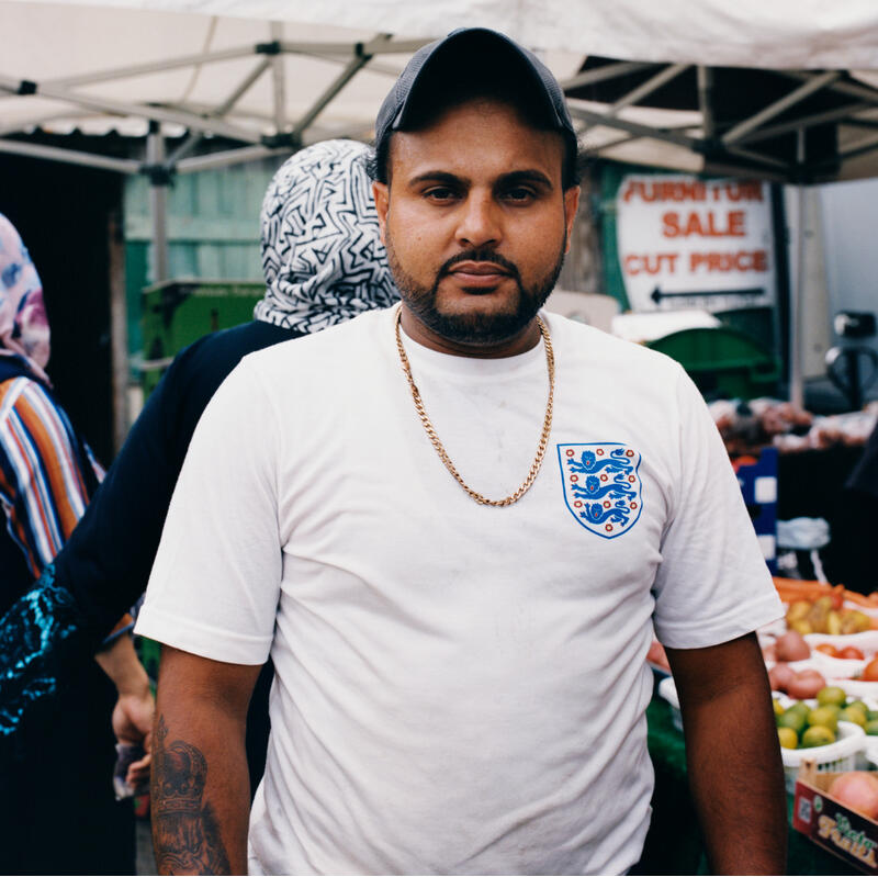 A man in a white shirt and black cap, with a chain necklace, stands under a market tent, with assorted produce in the background. Sign in background: "FURNITURE SALE CUT PRICE".