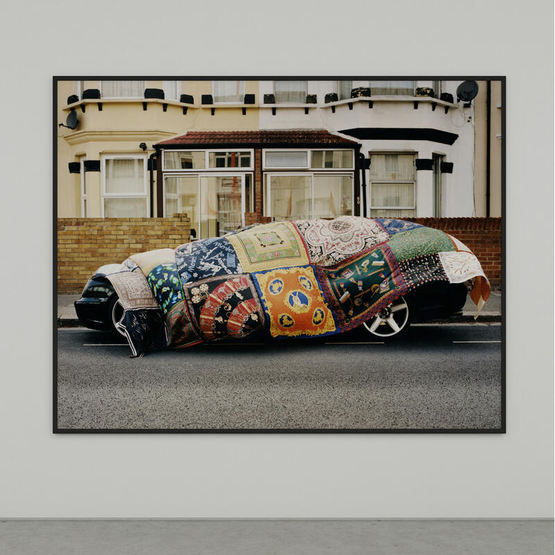 A quilt-covered car sits on a street, parked in front of a beige home with large windows and brick wall, as depicted in a framed photograph on a stark white gallery wall.