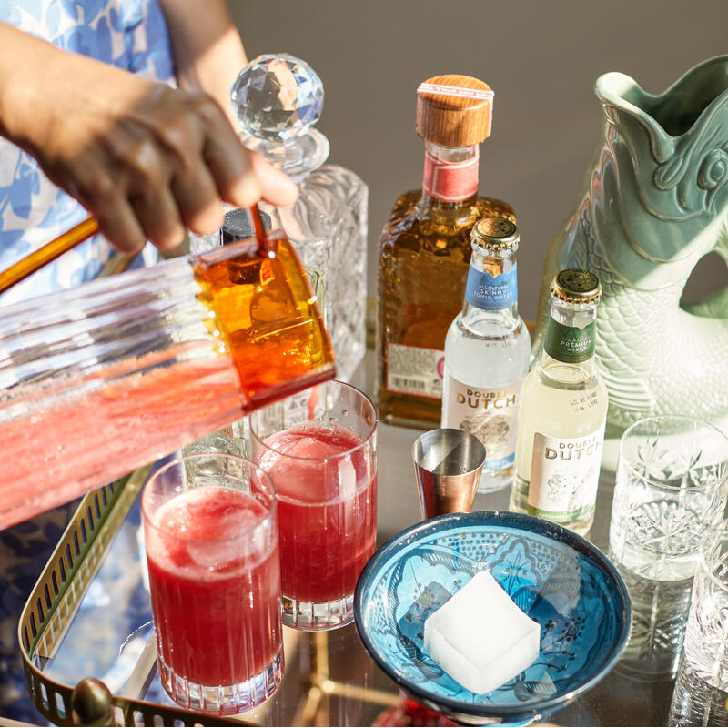 Hand pouring red liquid from an amber pitcher into glasses on a gold-trimmed bar cart, surrounded by various drink bottles, glasses, a blue bowl with an ice cube, and a green decorative vessel.