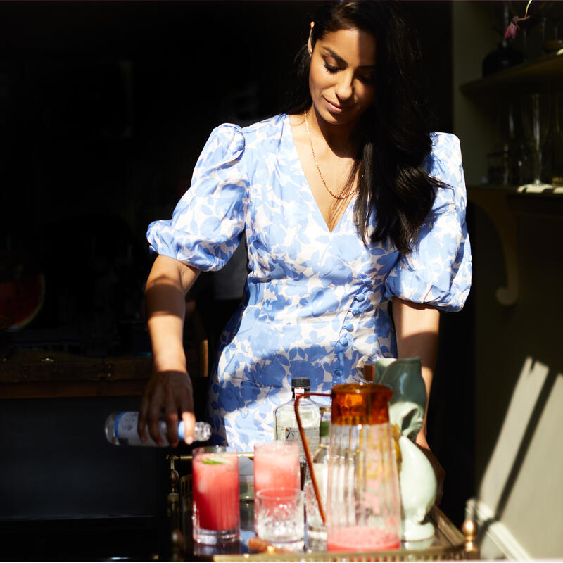 A woman in a floral blue dress pours a beverage into glasses on a tray in a warmly lit kitchen, with daylight streaming through a window above.