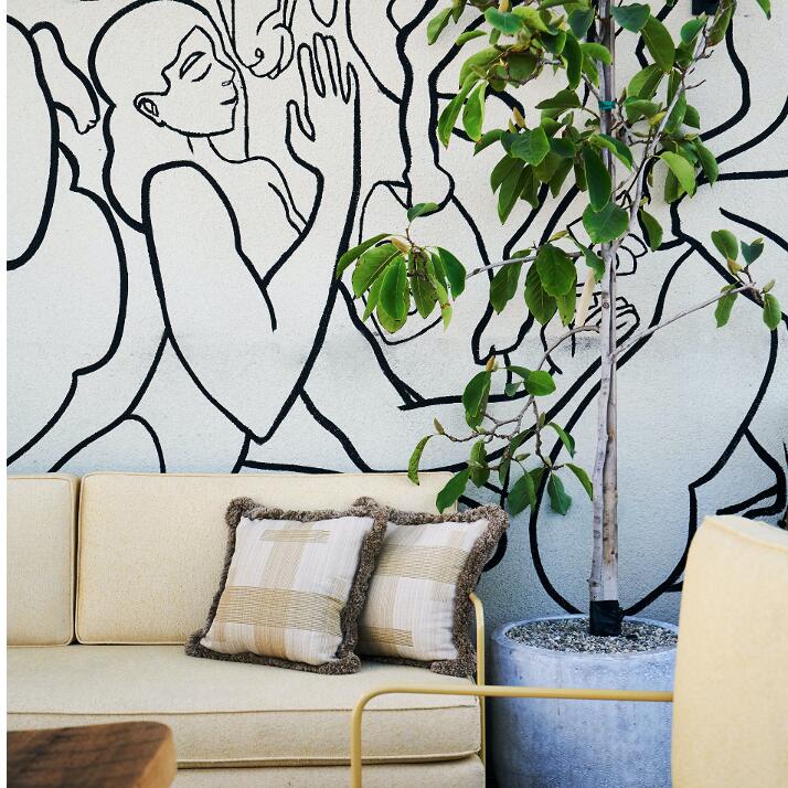 A beige sofa with two checkered pillows sits near a potted plant. A mural of a person and a horse is on the wall behind it, with a lit wall sconce above.