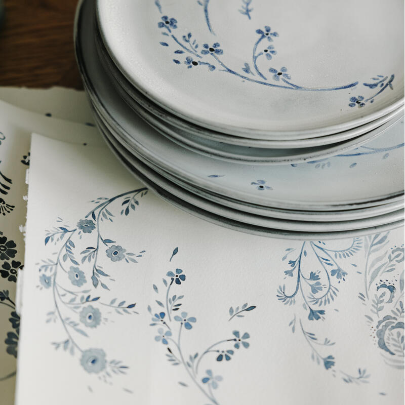 Stacked white plates adorned with blue floral designs lie on top of scattered papers featuring matching floral sketches on a wooden surface.