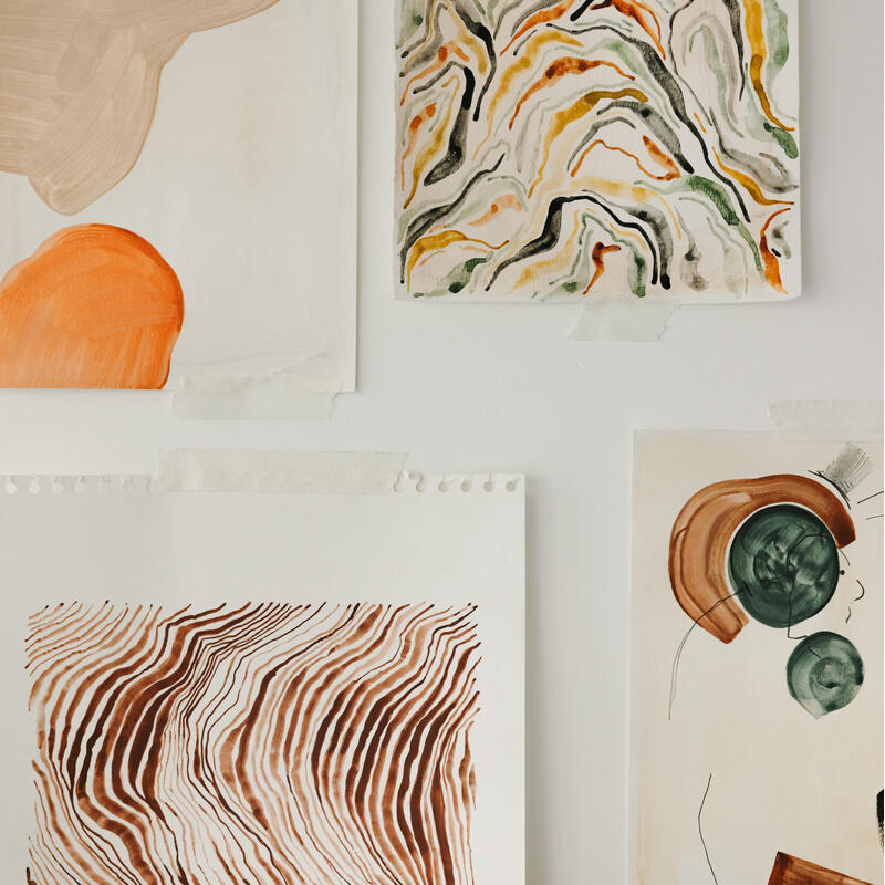 Four abstract paintings taped to a white wall, featuring organic shapes and swirling lines in earthy tones of brown, orange, green, and beige.