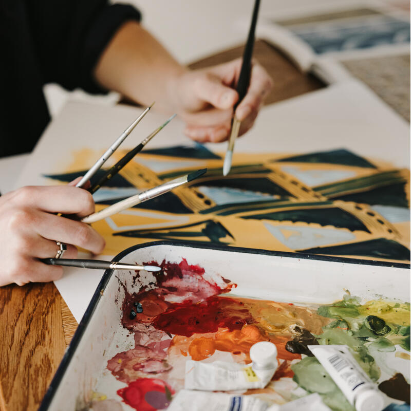 Several paintbrushes being held while painting a colorful abstract design on paper; nearby is a messy palette filled with various paint tubes and mixed colors.