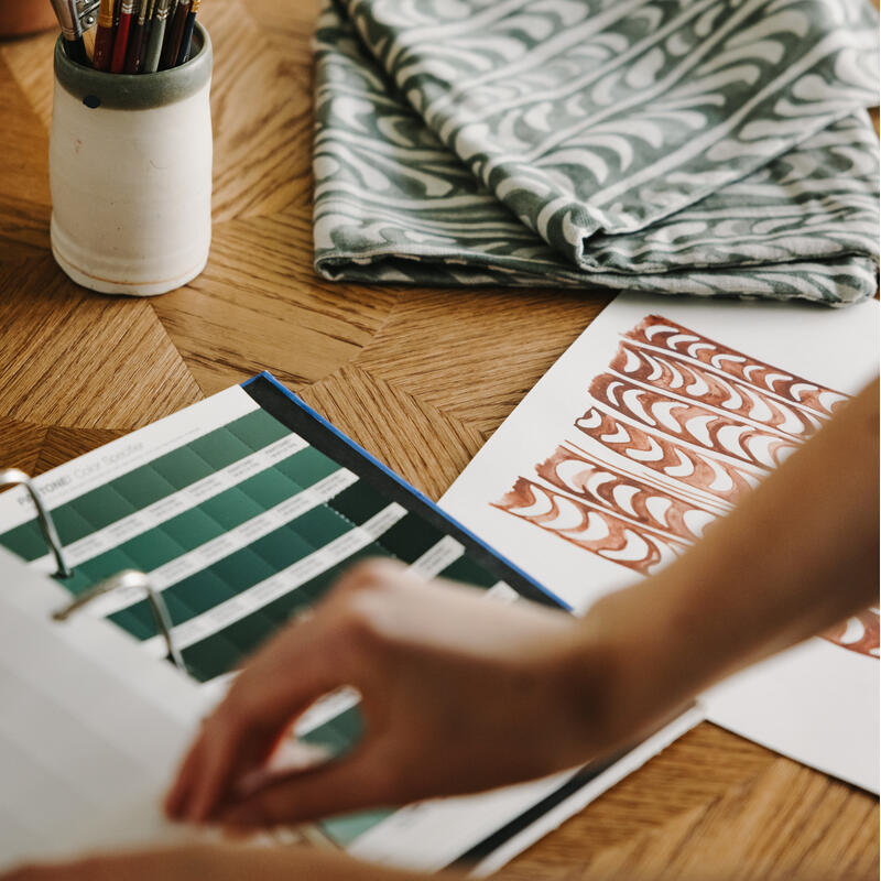 A collection of paintbrushes in a ceramic holder is accompanied by folded patterned fabric, color swatches, and patterned art on a wooden table, with a person reviewing a binder.