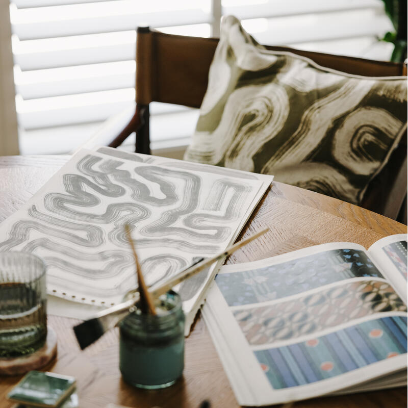 Sketches and art supplies sit on a wooden table in a sunlit room with a patterned pillow and a plant in the background.