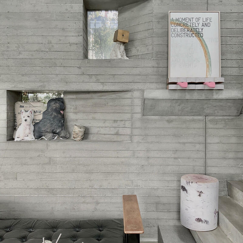 Art pieces and pillows are displayed on a concrete wall with an art piece with text: "A MOMENT OF LIFE CONCRETELY AND DELIBERATELY CONSTRUCTED". The area includes a black bench, modern sculpture, and concrete stairs.