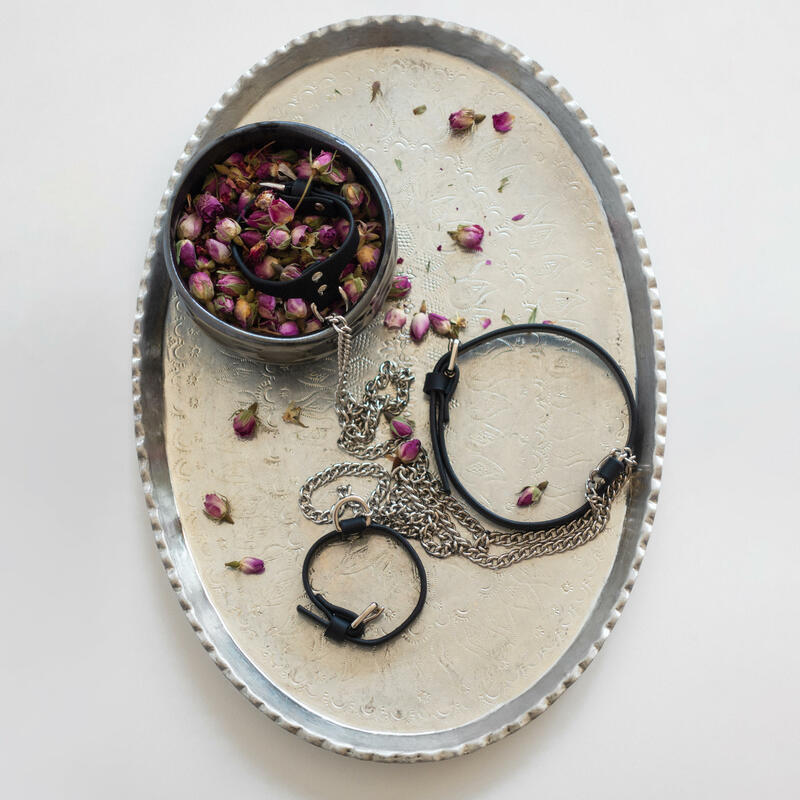 A metallic round tray holds black leather straps with attached silver chains, surrounded by scattered dried rosebuds, resting on a white surface.