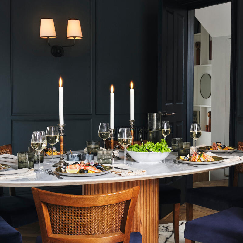 Wooden dining table set elegantly with plates of food, candles, and wine glasses; located in a modern room with dark walls, a colorful abstract painting, a sideboard, and a leafy plant.