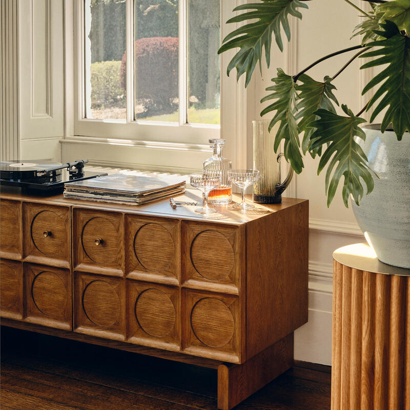 A wooden cabinet with a patterned front supports a turntable, vinyl records, and a drinks set near a large window overlooking a garden. Nearby, potted plants accent a well-lit, elegant room.