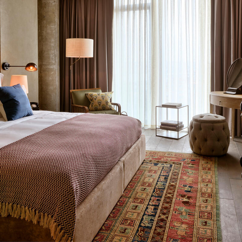 A neatly made bed with blue pillows and patterned blanket occupies a cozy, well-decorated bedroom featuring a wooden headboard, modern lamps, an armchair, sheer drapes, and a colorful rug.