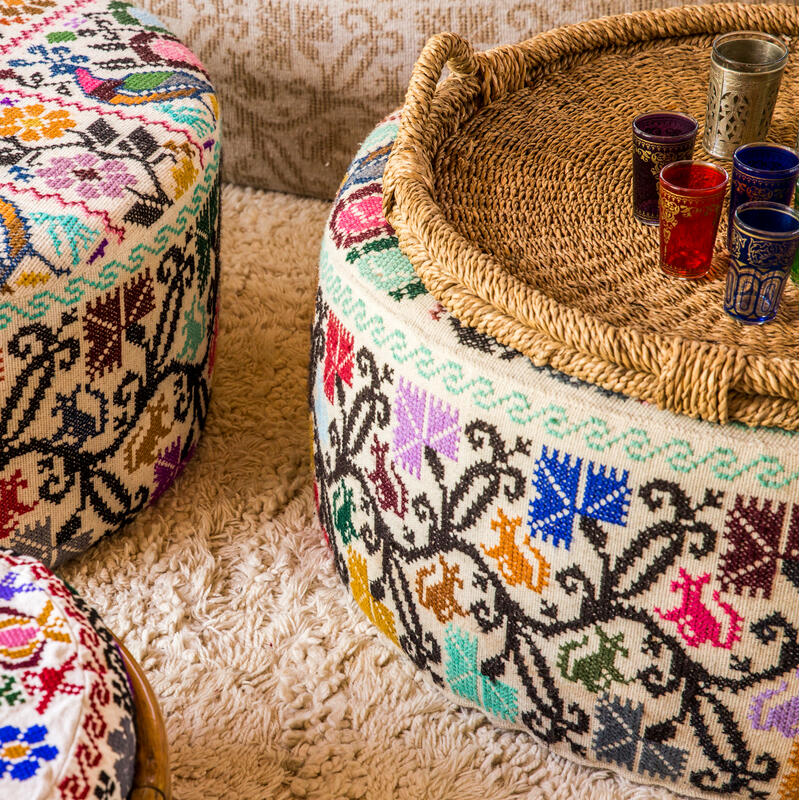 Three colorful, round, embroidered poufs with intricate patterns sit on a plush, cream carpet. One pouf holds a woven tray with several ornate, multicolored small cups. Background: patterned beige wallpaper.