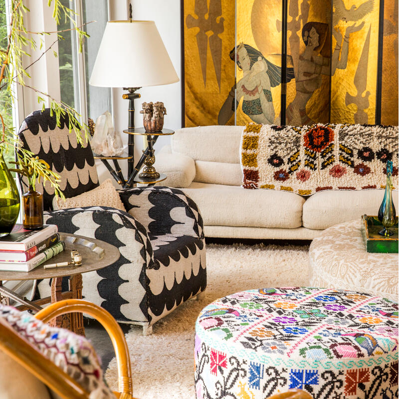 A cozy living room features a cream couch adorned with an ornate, multicolored blanket, surrounded by patterned chairs, a lamp on a side table, and various decorative items against a backdrop of artistic wall panels.