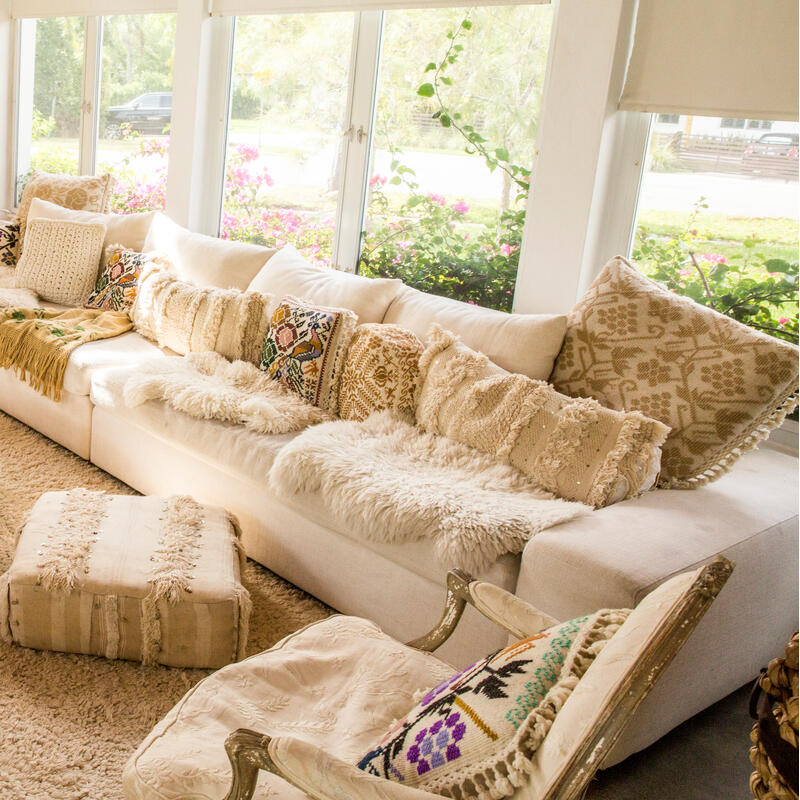 A cozy, bright living room with large windows, a white L-shaped sofa filled with patterned cushions, a round ottoman with a green tray and decorative objects, and various artworks and plants.