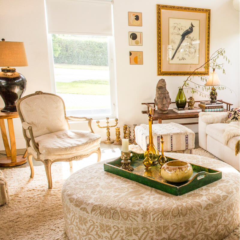 A white, cozy living room features a large sectional sofa with plush pillows, a patterned ottoman with decor on a tray, and floor-to-ceiling windows showcasing a lush garden.