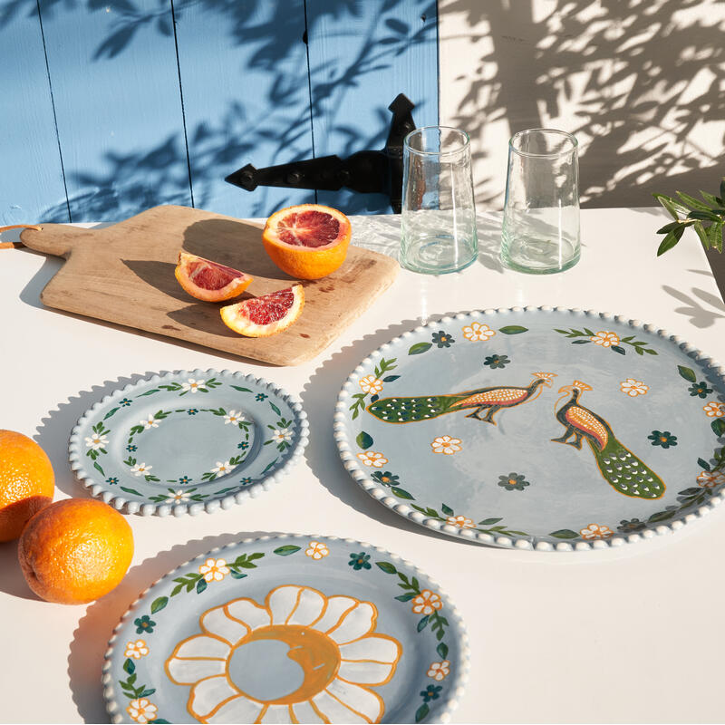 Ceramic plates decorated with flowers and birds rest on a table. Nearby, a wooden cutting board holds a sliced blood orange. Glass tumblers and whole oranges are present, all against a blue wooden wall with tree shadow patterns.