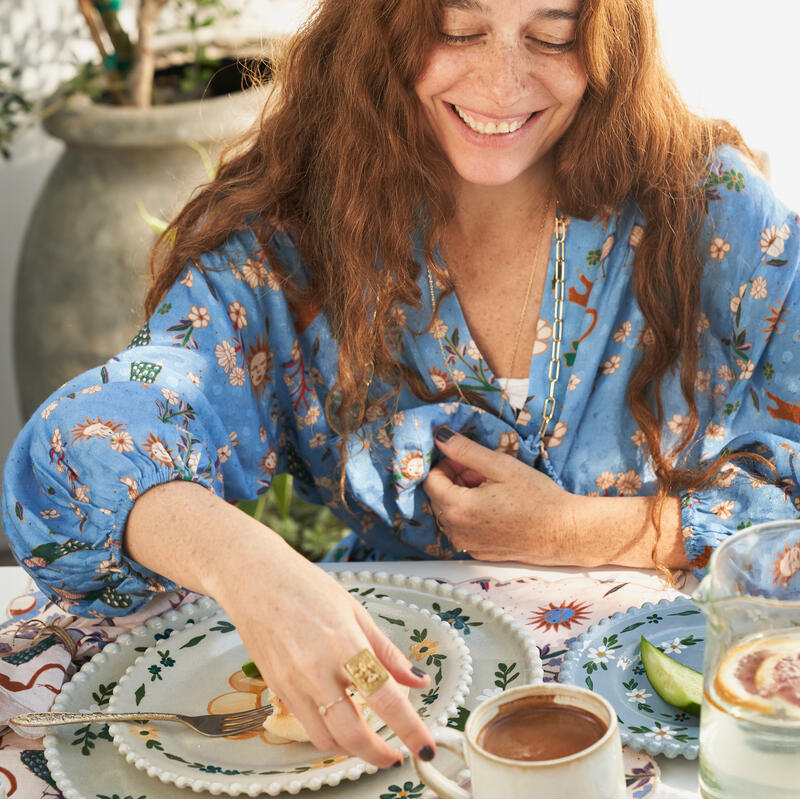 A smiling woman wearing a blue floral blouse reaches for a white mug while sitting at a table adorned with decorative plates, orange slices, and a pitcher of citrus water outdoors.