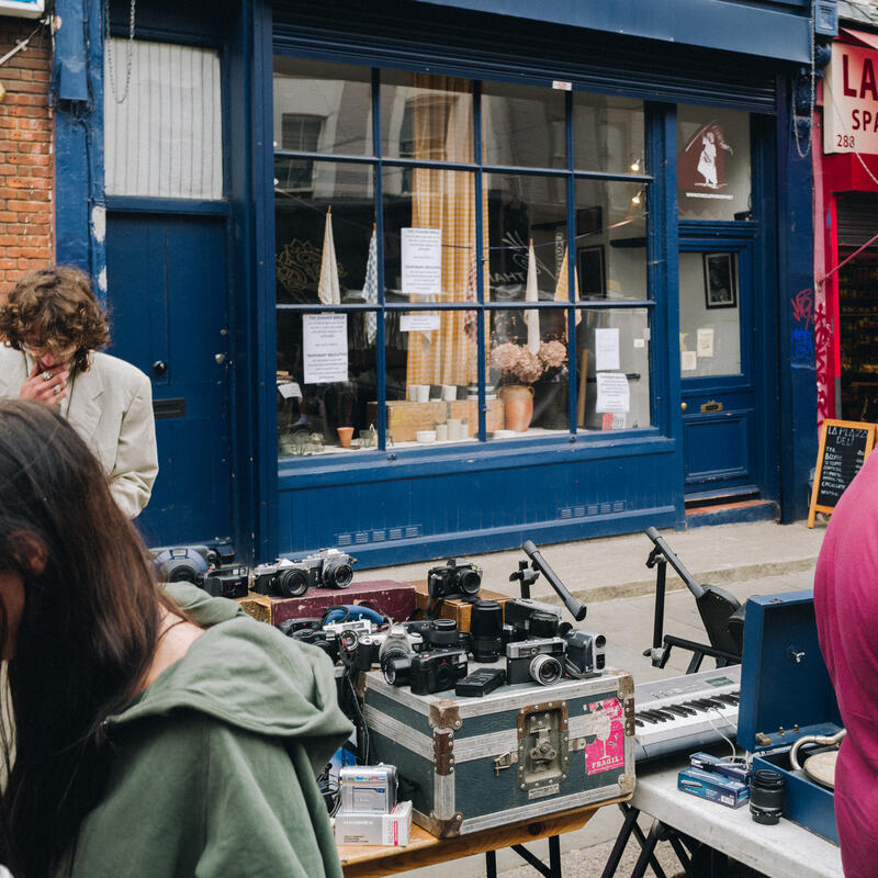 A street market table displays various vintage cameras and equipment outside a blue storefront. Two people inspect items while another store entrance with a red awning is partially visible.