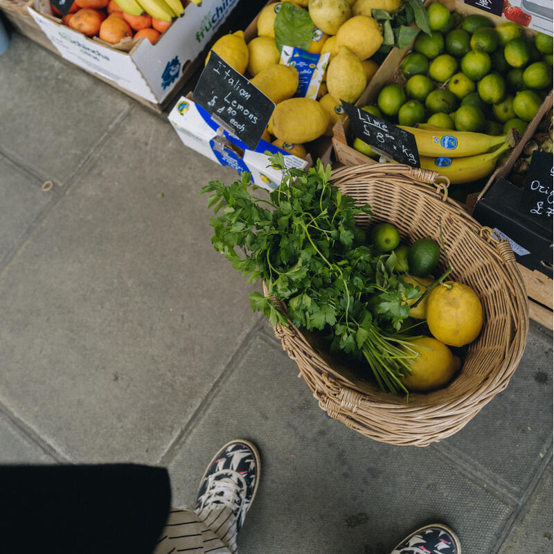 Various fruits and vegetables, including bananas, citrus fruits, and greens, are displayed in baskets and crates on a sidewalk, with text labeling them, viewed from a person standing above. The text seen reads:

1) "Set 'L' pec. kg."
2) "Set 'G' pec. kg."