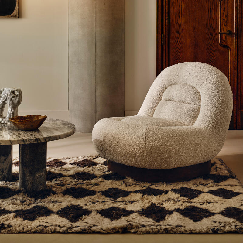 A plush white armchair sits on a black-and-white geometric rug beside a round marble table in a contemporary living room, featuring a wooden door, concrete pillar, and modern light fixture.