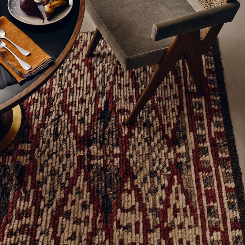 A partially visible wooden dining table with fruit on a plate, an upholstered chair, and a colorful woven rug featuring red, brown, and beige geometric patterns in a modern dining room.