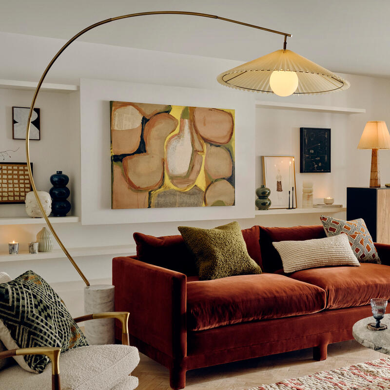 A brown velvet sofa with decorative cushions sits in a cozy living room, beneath a unique arc lamp. Abstract art, various ornaments, and a textured rug enhance the sophisticated environment.