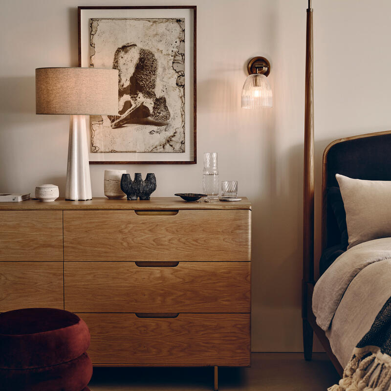 A wooden dresser with decorative items and a lamp on top stands next to a bed, with a framed painting above and a wall light fixture illuminating it in a cozy bedroom.