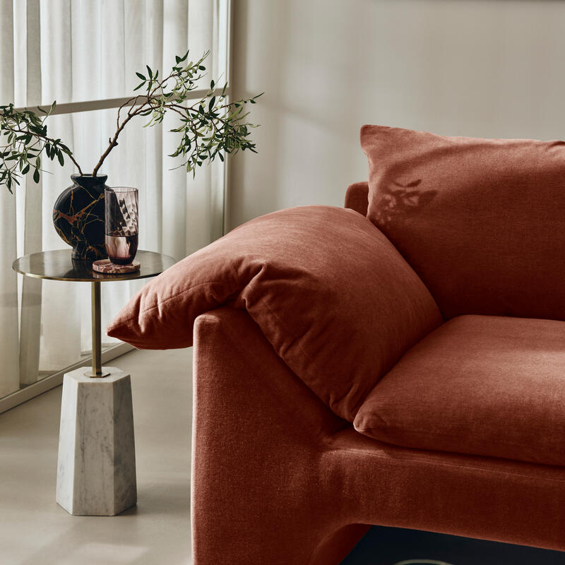 A soft, rust-colored sofa sits in a modern living room, accompanied by a marble side table holding a black vase with greenery and a glass. Sheer curtains filter natural light.