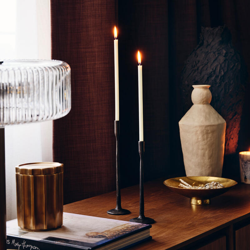 Two tall candlesticks with lit candles stand on a wooden table next to a brass container, a book, a lamp, a beige vase, and a brass bowl, with dark curtains in the background.