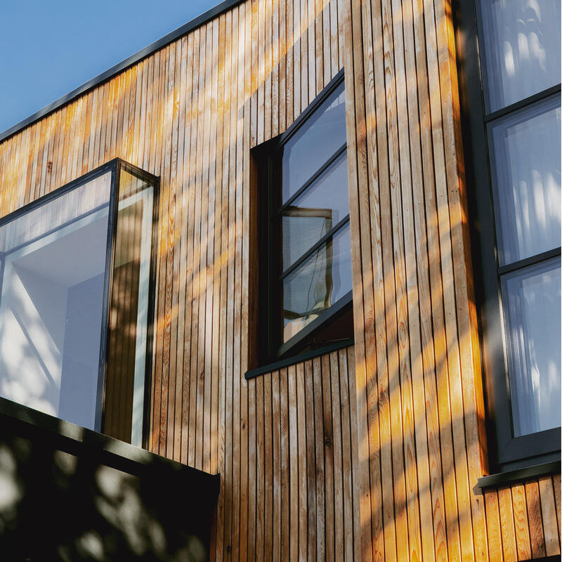 Wood-clad modern building features large glass windows, casting shadows from nearby trees, set against a clear blue sky.