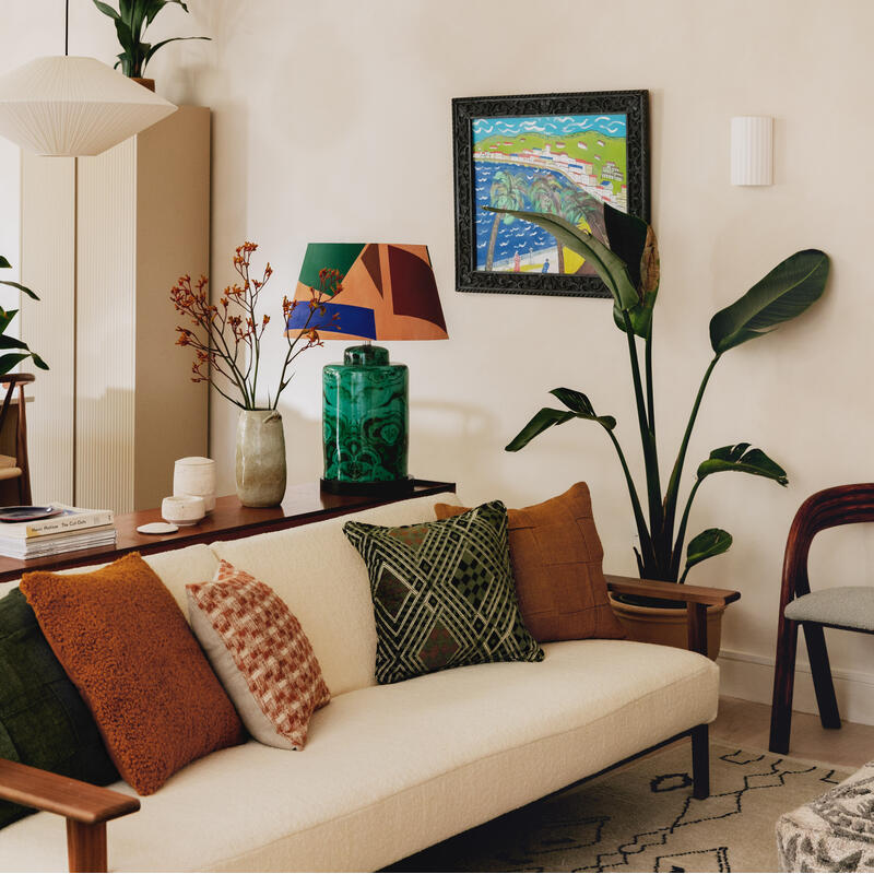 A cozy living room scene featuring a white sofa adorned with colorful pillows; a wall with a vibrant painting, plants, books, and a green decorative jar in soft lighting.