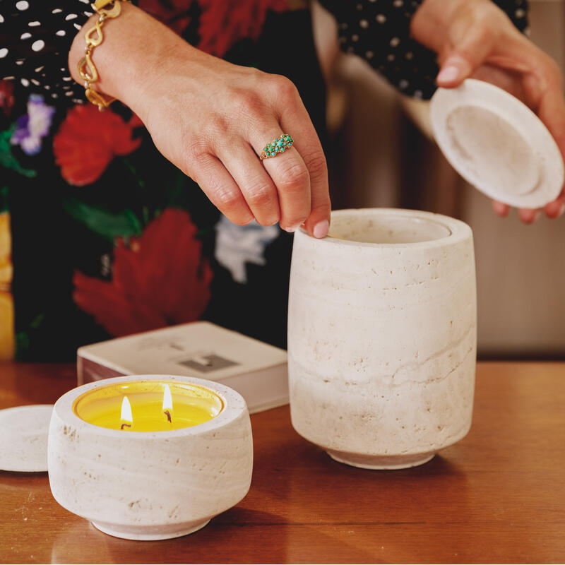 A person with a green gemstone ring and gold bracelet is lifting the lid off a white ceramic container beside a lit yellow candle on a wooden table in a cozy room.