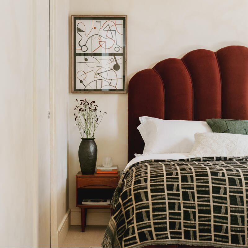 A cozy bedroom features a bed with a burgundy, scalloped headboard, and patterned bedding, a black vase with flowers on a nightstand, and abstract artwork on the wall.