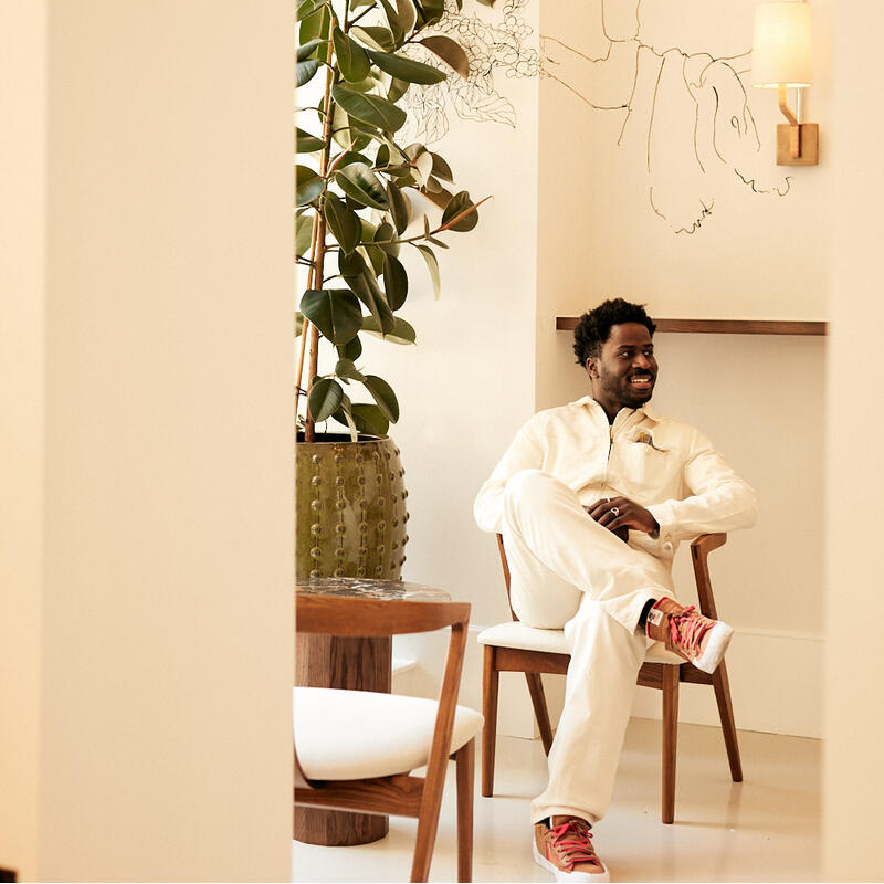 A man in white clothing and red sneakers sits on a wooden chair, smiling, next to a large potted plant in a bright, beige room with minimalist decor and wall art.