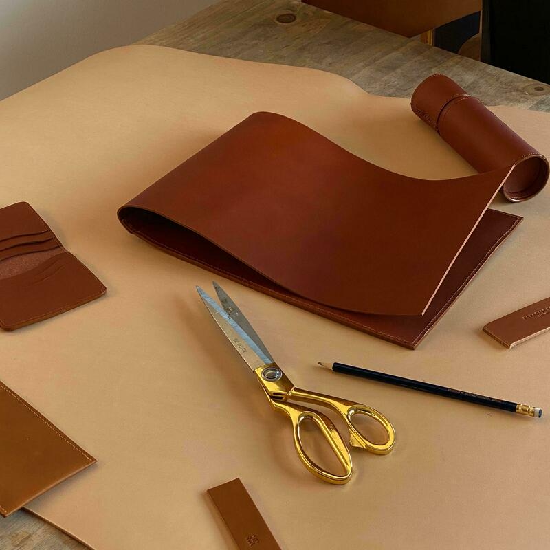 Golden scissors, a pencil, and strips of brown leather lie on a wooden table, beside larger leather pieces, in a workshop setting.