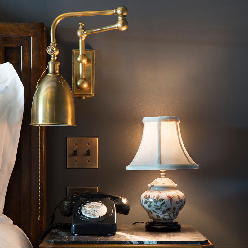 A brass wall-mounted lamp, black rotary telephone, and floral-patterned table lamp rest on a wooden nightstand beside a bed with white sheets, set against a dark gray wall with brass light switches.