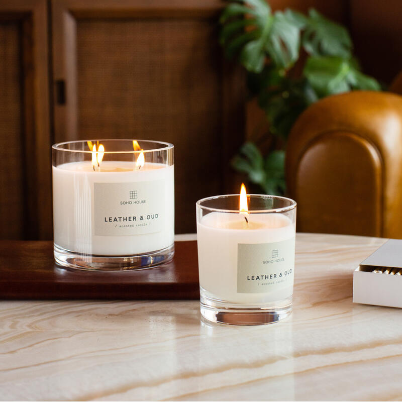 Lit white candles labeled "SOHO HOUSE LEATHER & OUD scented candle" rest on a wooden and marble surface in a cozy room with warm brown tones, a plant, and a leather chair.